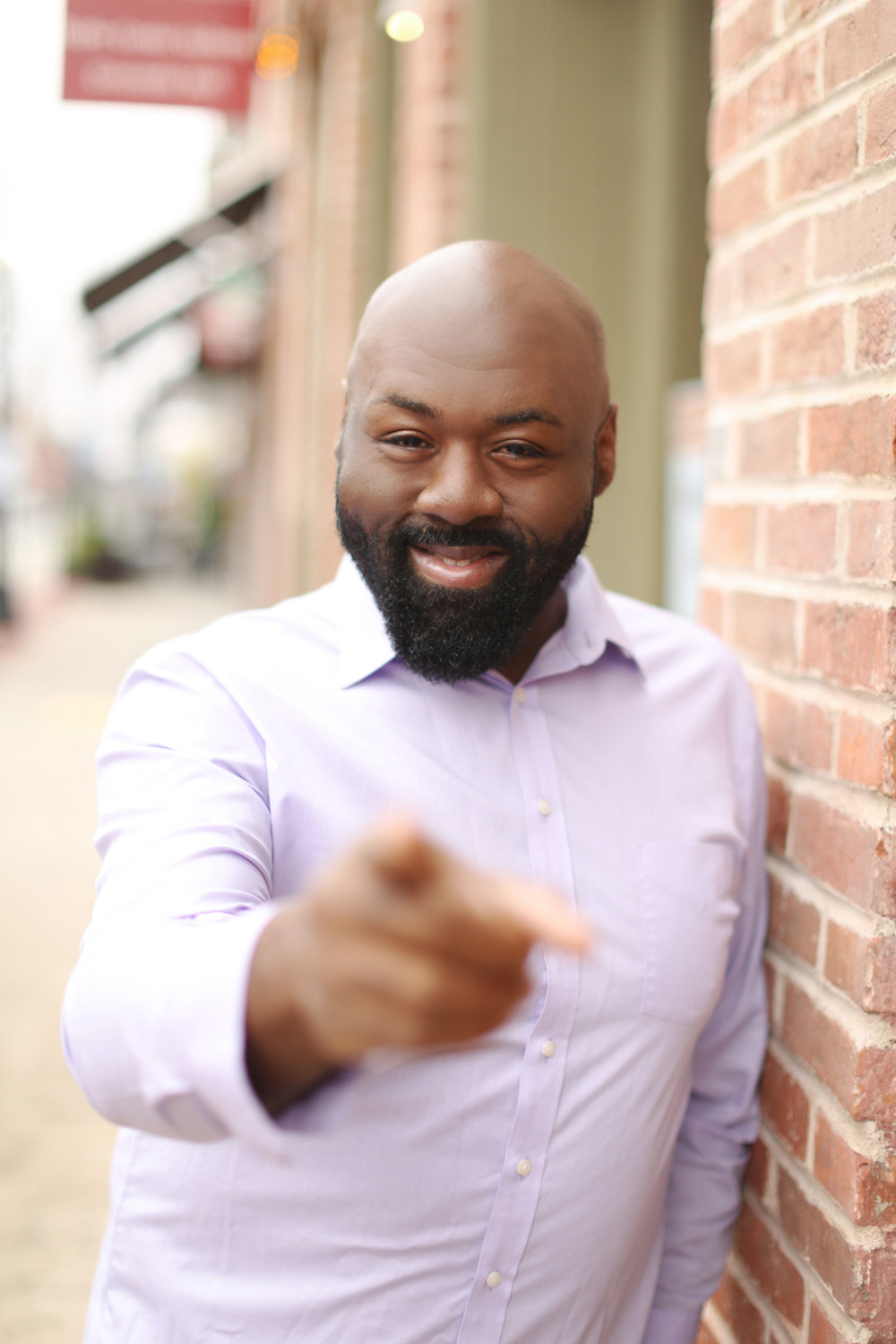 Bald bearded man in a lavender shirt leaning on a red brick wall, pointing at the camera