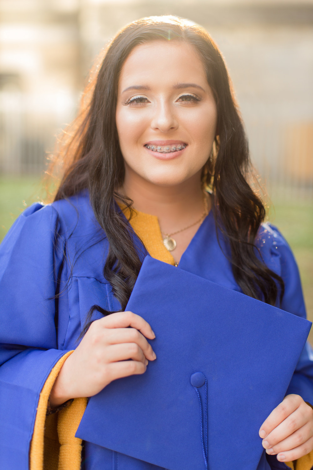 Graduate in a blue gown holding her cap, backlit by golden light on campus